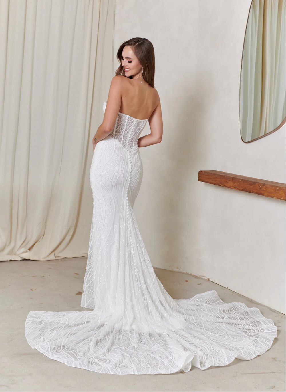 Woman in a white lace wedding dress standing in a room with a mirror and draped fabric.