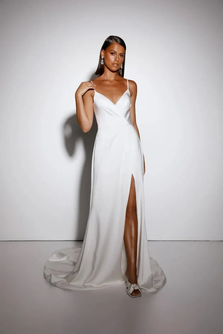 A woman modeling a floor-length white gown with thin straps, a draped neckline, and a high front slit, standing against a plain light gray studio background.