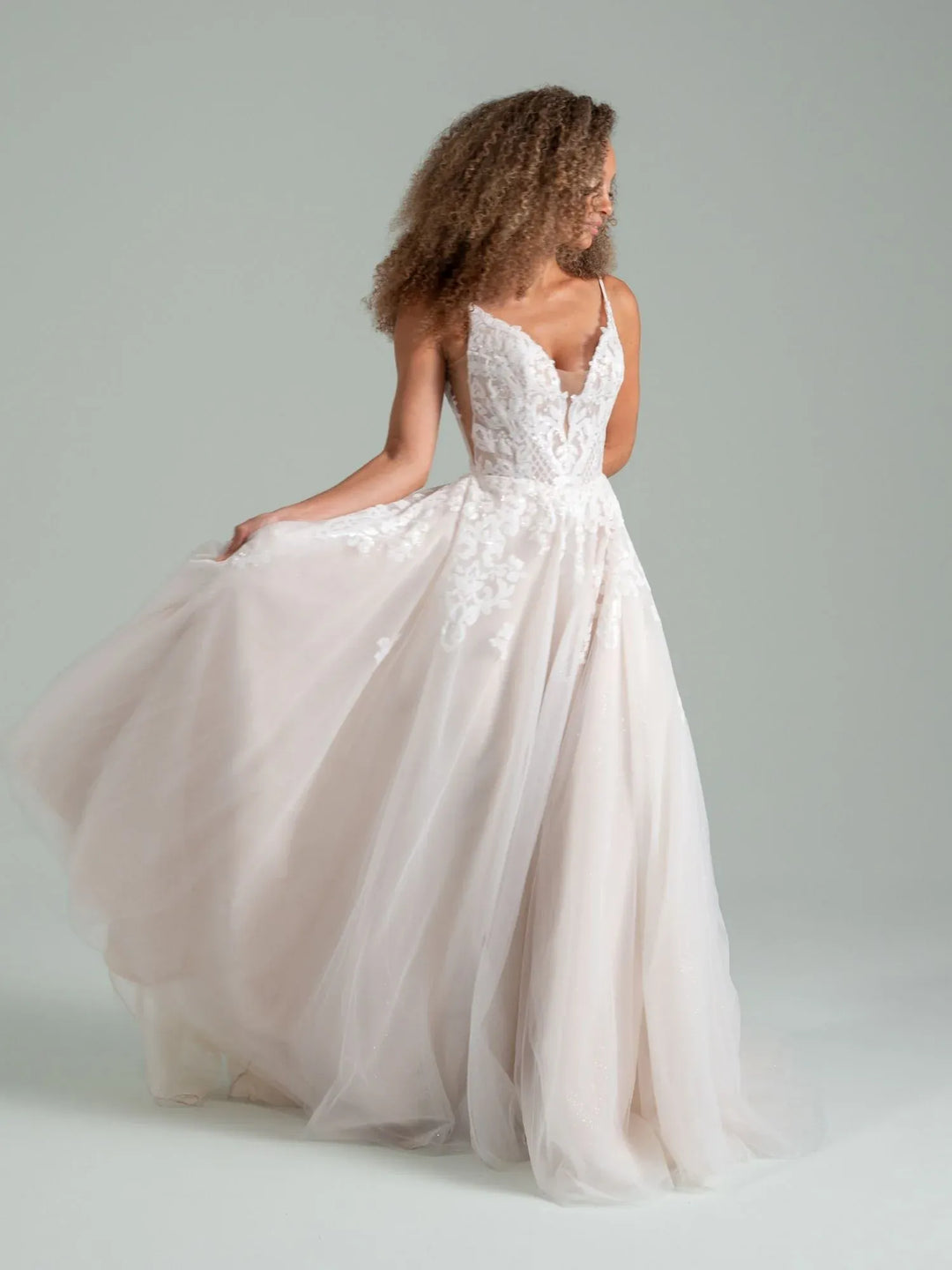 A woman modeling a white A-line wedding gown with lace bodice, spaghetti straps, and a full tulle skirt against a light gray studio background.