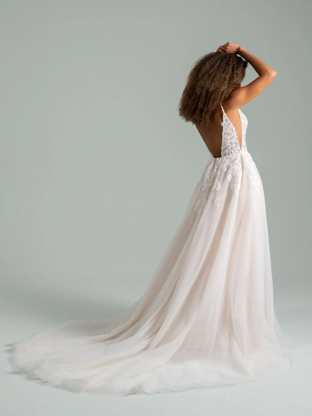 A woman wearing a white wedding dress with lace detailing and a long flowing tulle train, photographed from the back against a plain light gray studio background.