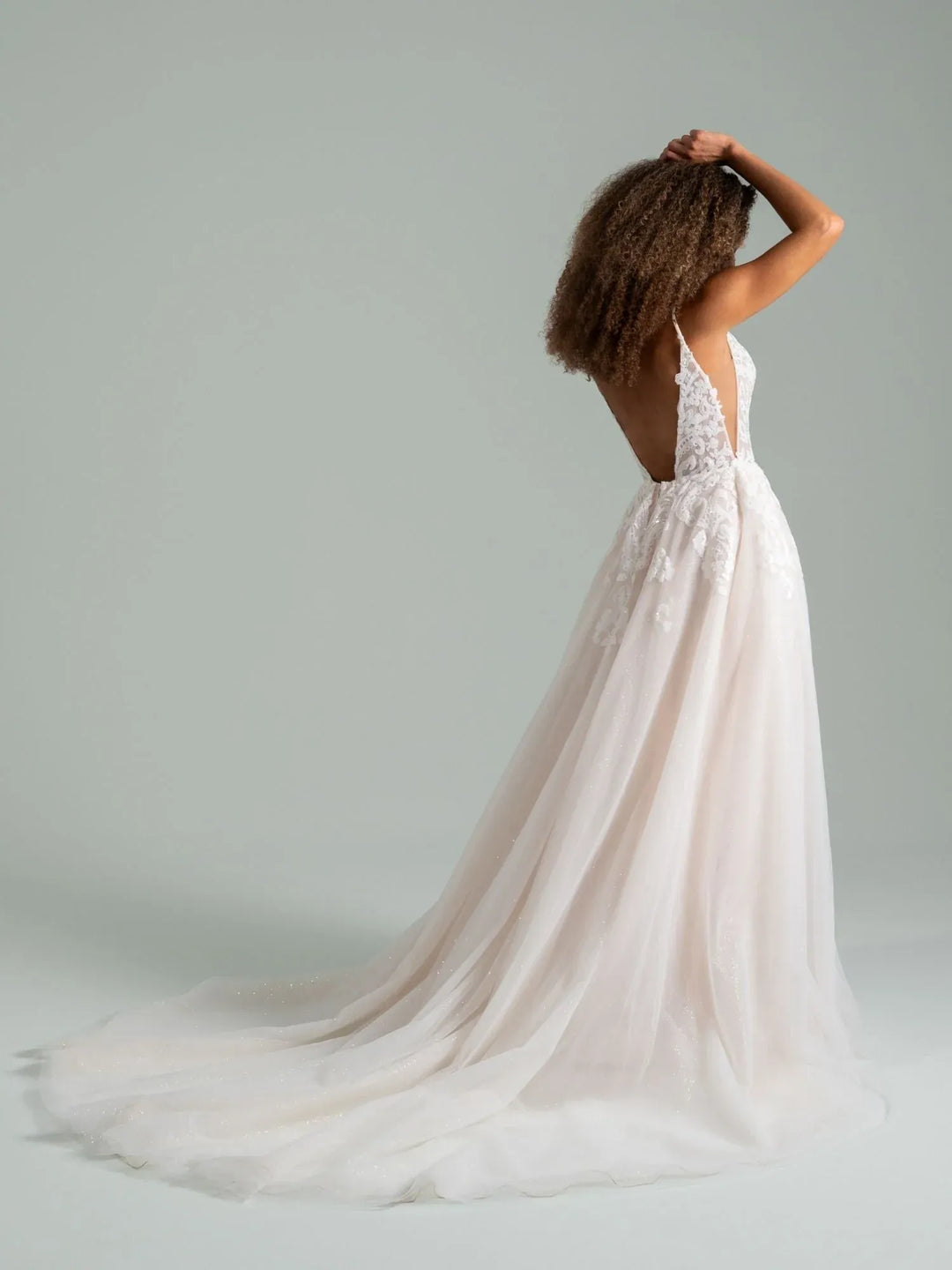 A woman wearing a white wedding dress with lace detailing and a long flowing tulle train, photographed from the back against a plain light gray studio background.