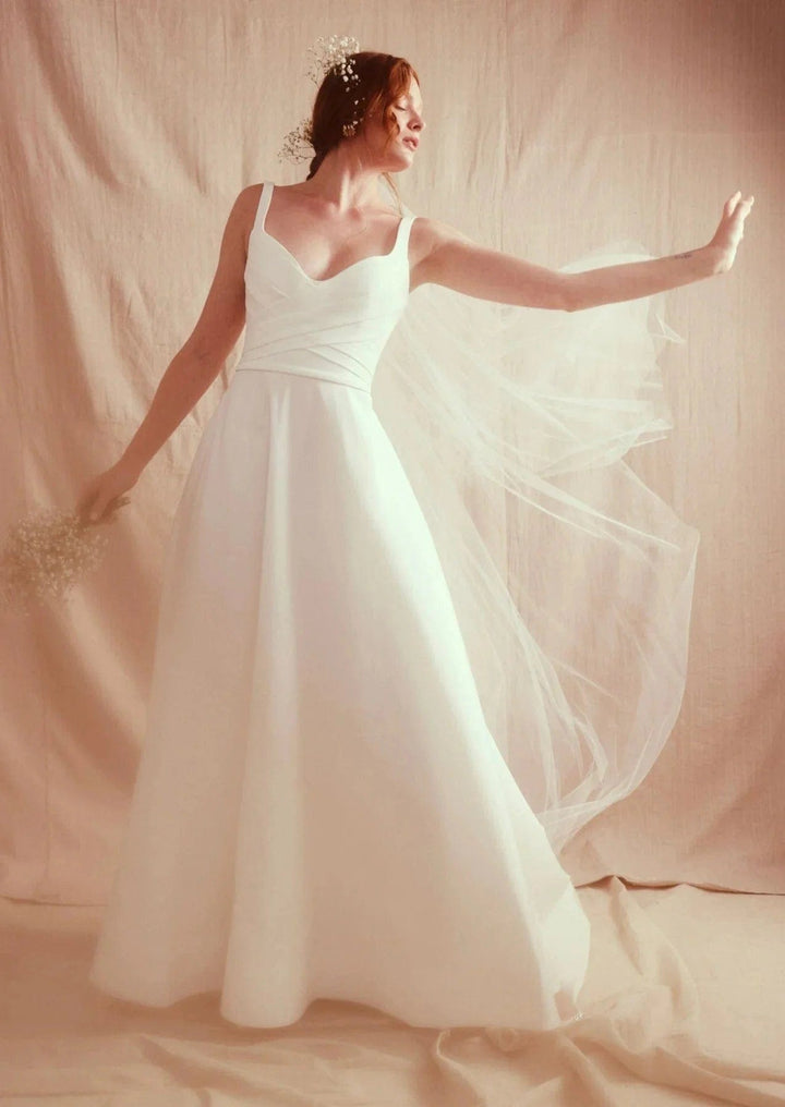 Woman in a white wedding dress with a flowing veil against a soft pink background