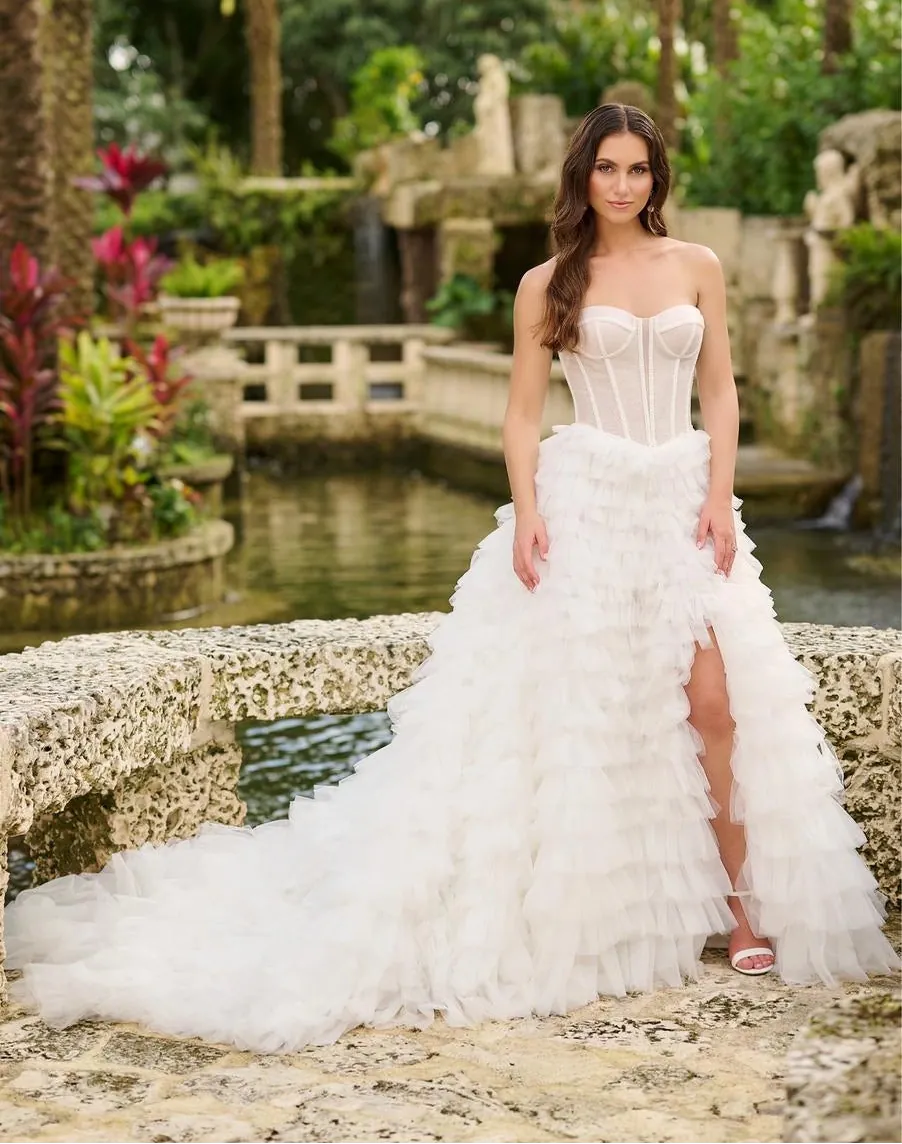 A woman wearing a strapless white wedding gown with a structured corset-style bodice and a voluminous ruffled overskirt, standing outdoors on a stone bridge with water and tropical greenery in the background.