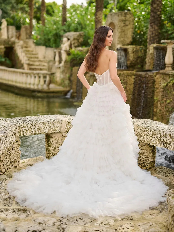 A woman wearing a white strapless wedding gown with a structured corset-style bodice and a voluminous layered tulle skirt, standing on a stone bridge in a garden setting.
