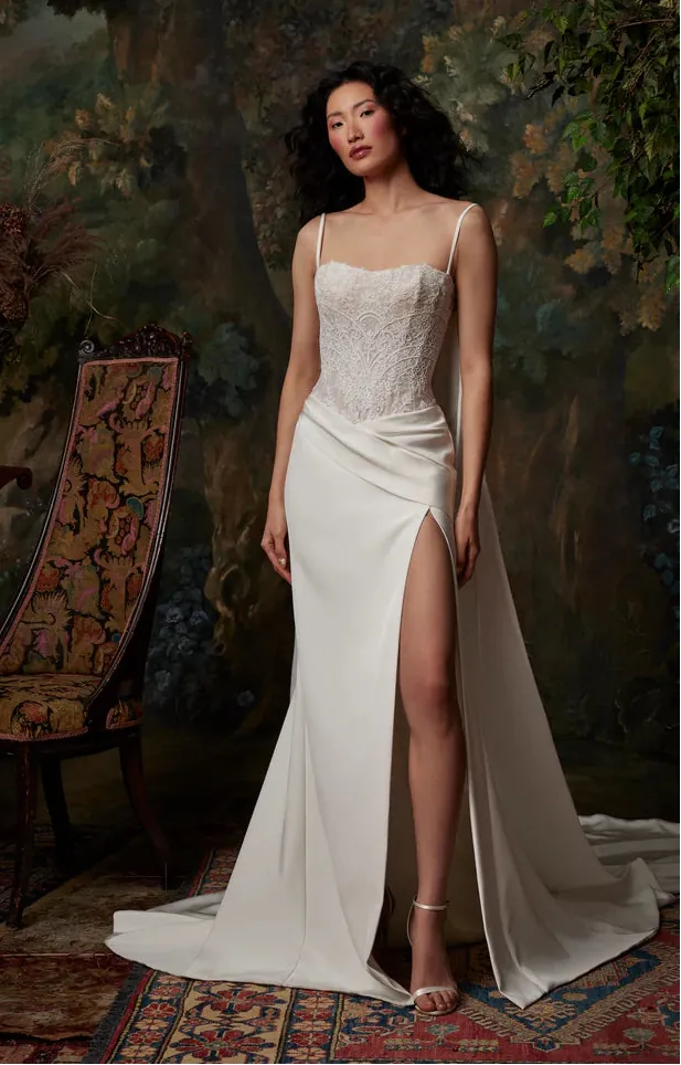 A woman models a sleeveless ivory wedding gown with a lace-embroidered bodice, thin straps, a draped skirt, and a high thigh slit. She stands in a richly decorated room with a large painted backdrop, an ornate chair, and patterned rugs.
