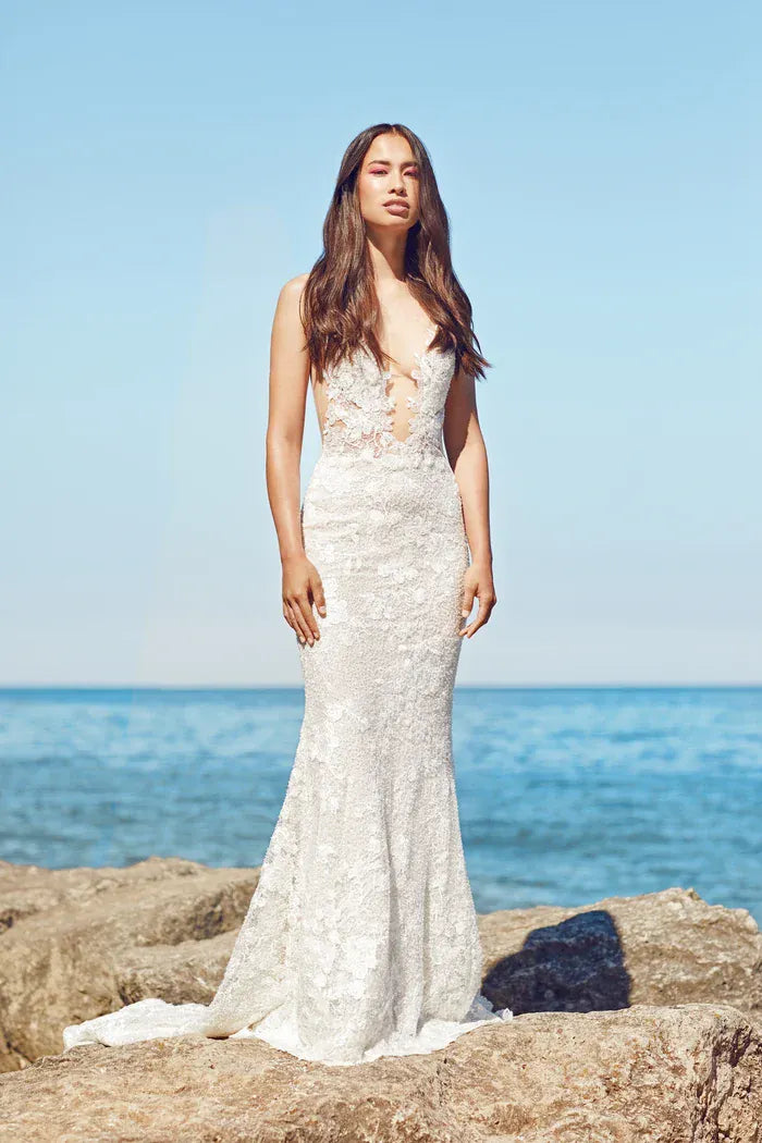 A woman wearing a sleeveless lace mermaid-style wedding gown stands on rocks by the ocean under a clear blue sky.