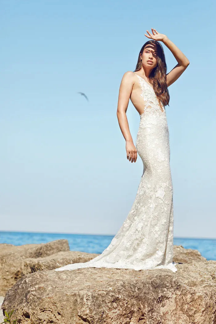 A woman wearing a sleeveless lace wedding gown stands on large rocks by the ocean under a clear blue sky, with one hand raised to shield her eyes from the sun.