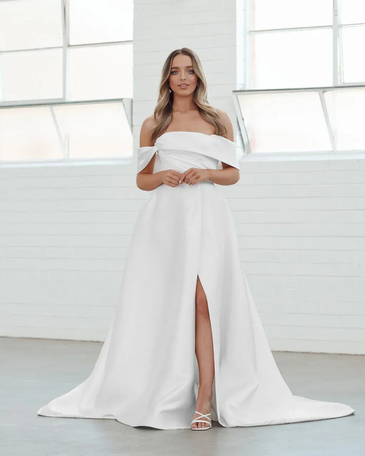 A woman modeling a white off-the-shoulder maxi dress with a high thigh slit and a slight train, standing in a bright white studio with large windows.