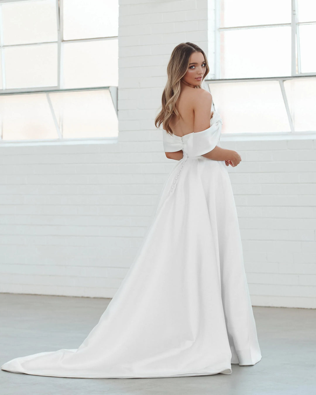 A woman wearing a white off-the-shoulder wedding dress with a long train, standing in a bright white room with large windows.