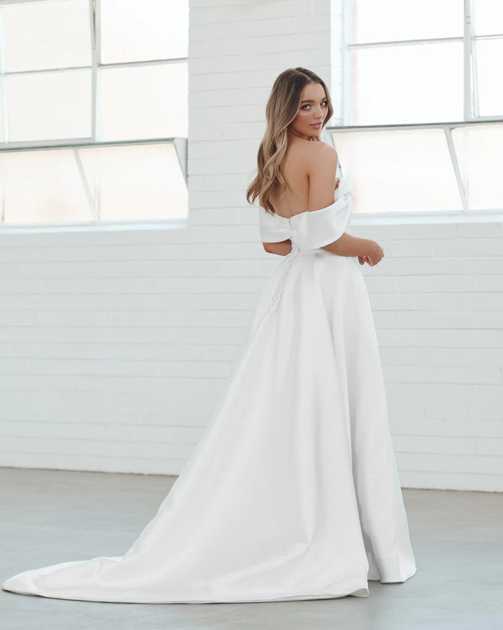 A woman wearing a white off-the-shoulder wedding dress with a long train, standing in a bright white room with large windows.