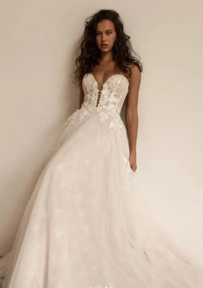 A woman modeling a strapless white wedding gown with a deep V neckline, lace applique bodice, and a voluminous tulle skirt against a plain light beige wall.