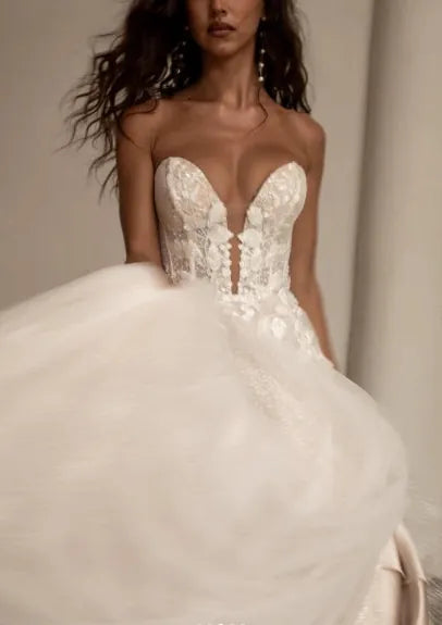 A woman modeling a strapless white lace wedding gown with a sweetheart neckline, lace-up front, and a voluminous tulle skirt in a softly lit indoor setting.