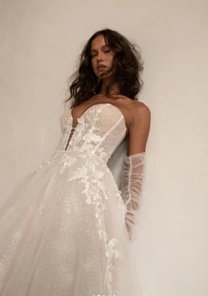 A woman wearing a strapless white lace wedding gown with a full tulle skirt and sheer long gloves, posed against a plain light beige wall.
