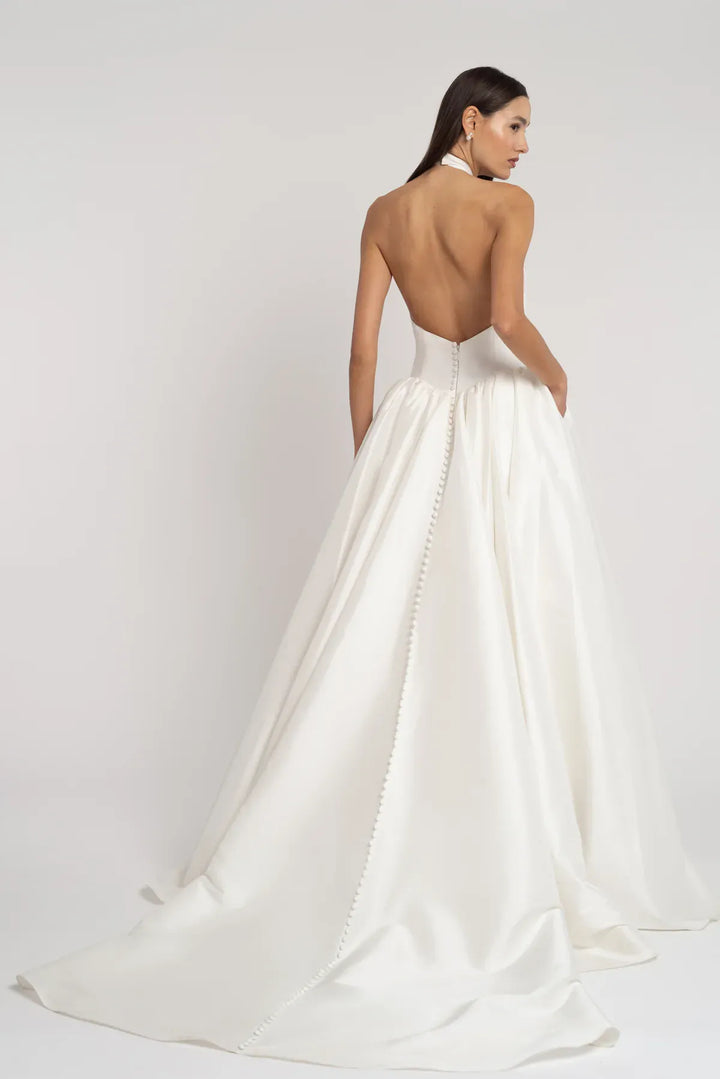 A woman modeling a white bridal gown from the back, showing an open back, waistband details, and a full satin train against a light gray studio background.