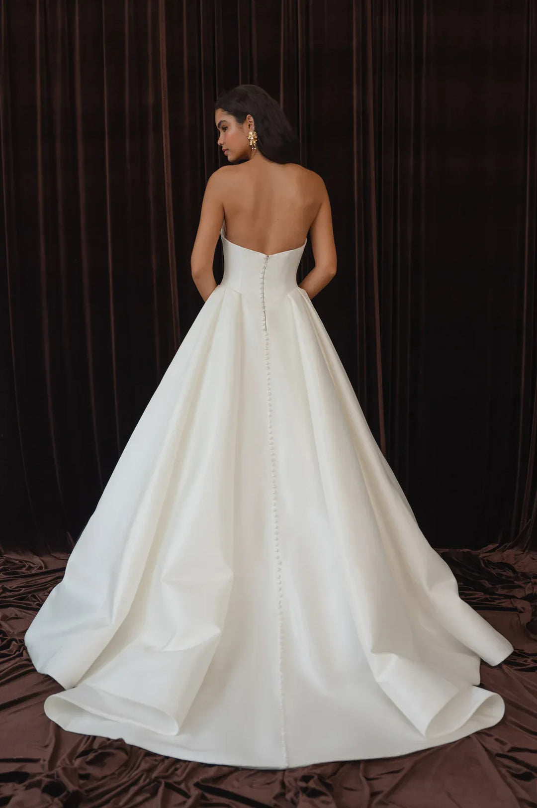 A woman wearing a strapless white ball gown with a structured bodice and full skirt, photographed from the back against a dark brown velvet curtain backdrop.