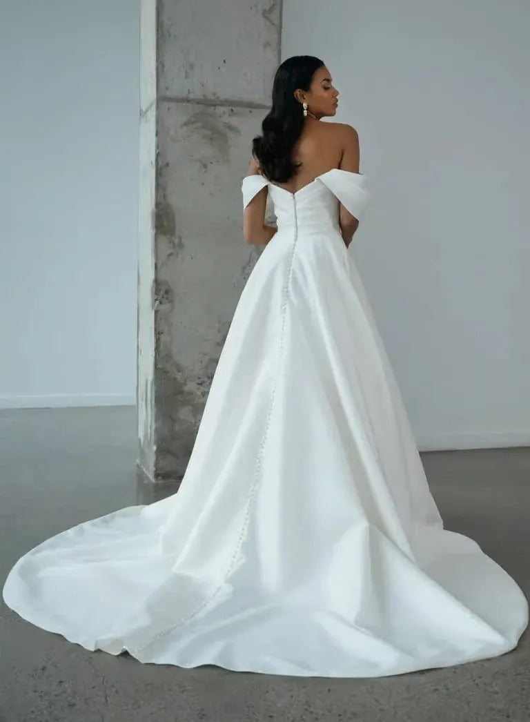 A woman wearing an off-the-shoulder white wedding gown with a structured bodice and a long train, photographed in a minimalist indoor setting with a concrete pillar.