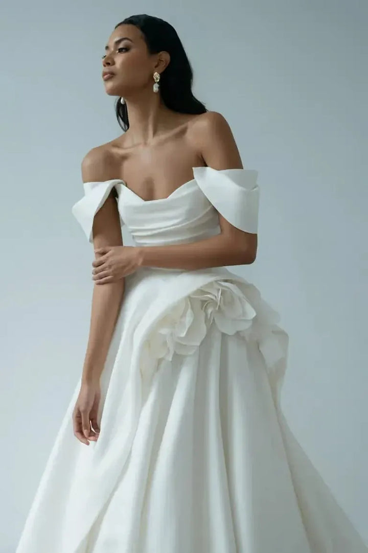 A woman modeling an off-the-shoulder white wedding gown with a structured sweetheart neckline, dramatic draped sleeves, and a voluminous ruffled skirt against a light gray studio background.