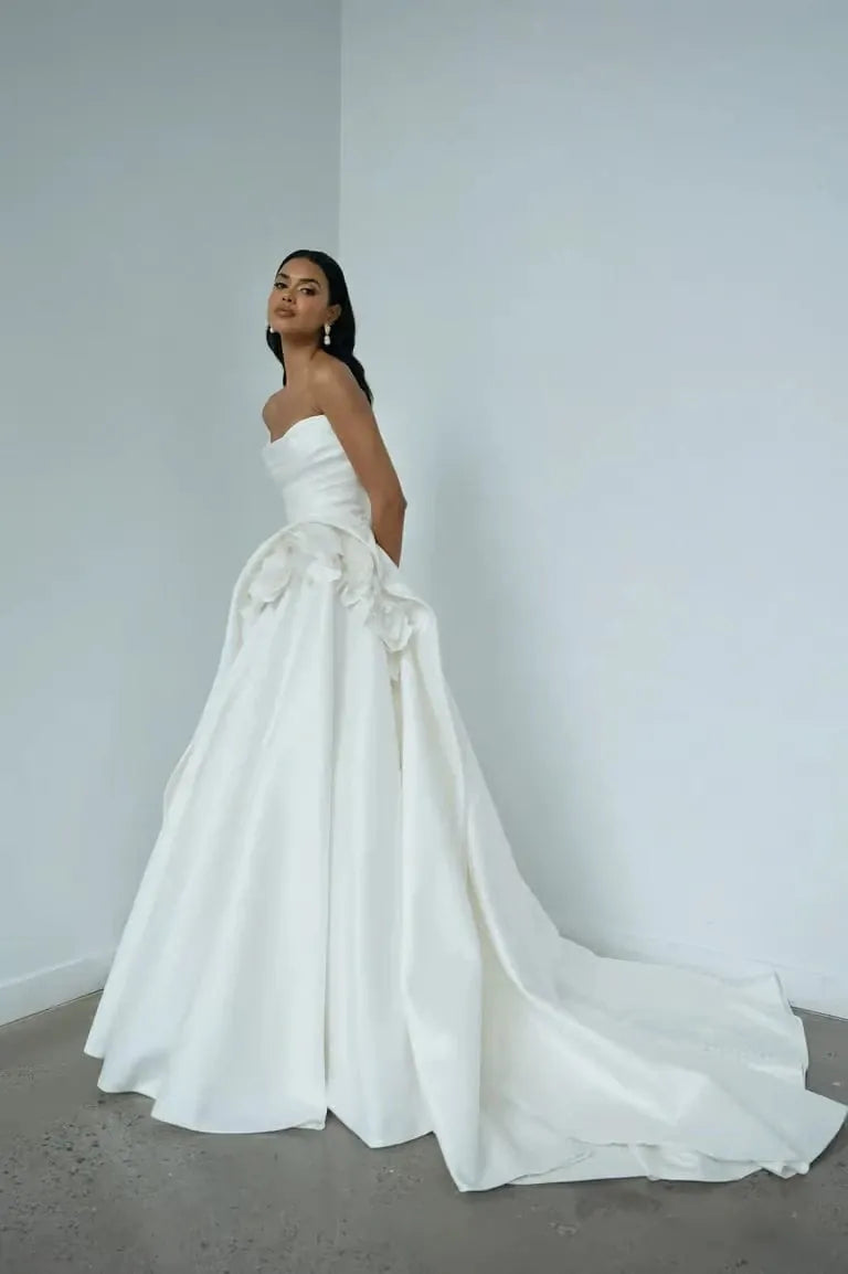 A woman modeling a strapless white wedding gown with a voluminous skirt, large floral applique at the waist, and a long train, standing in a minimalist white room with a gray floor.