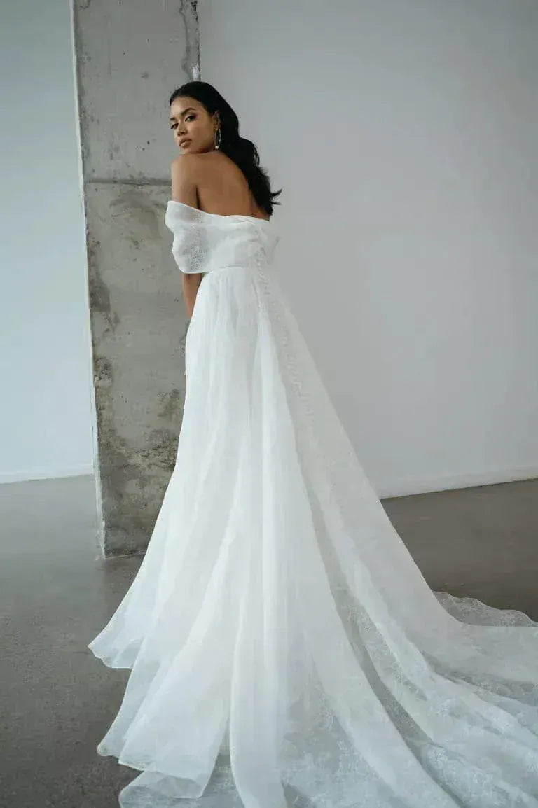 Woman in a white off-shoulder gown standing against a plain wall.