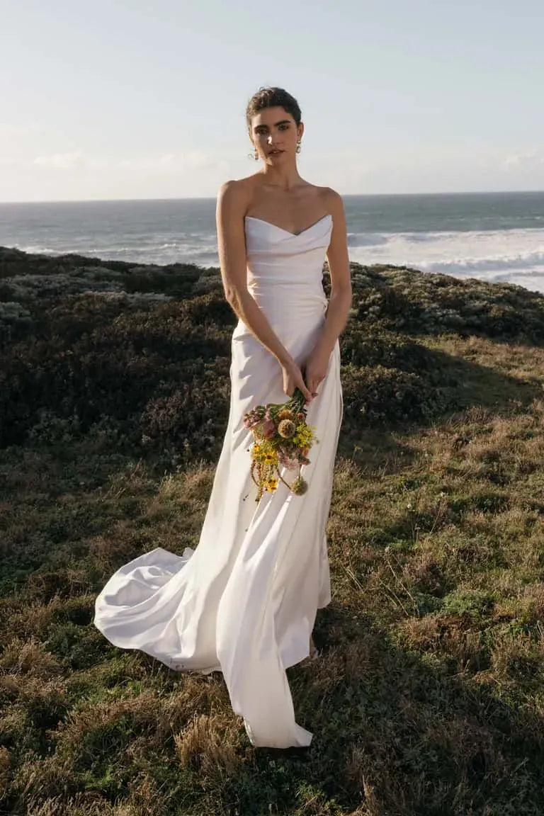 Woman in a white wedding dress holding a bouquet by the ocean.