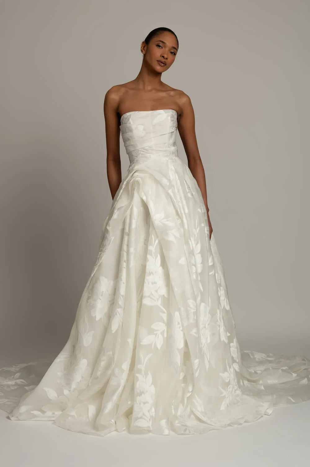 A woman modeling a strapless white bridal gown with a structured bodice and a full, flowing skirt featuring tonal floral jacquard texture, photographed against a plain light gray studio background.