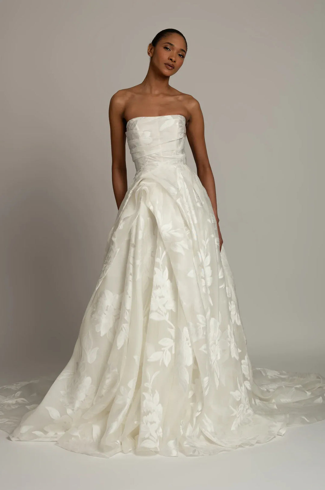 A woman modeling a strapless white bridal gown with a structured bodice and a full, flowing skirt featuring tonal floral jacquard texture, photographed against a plain light gray studio background.