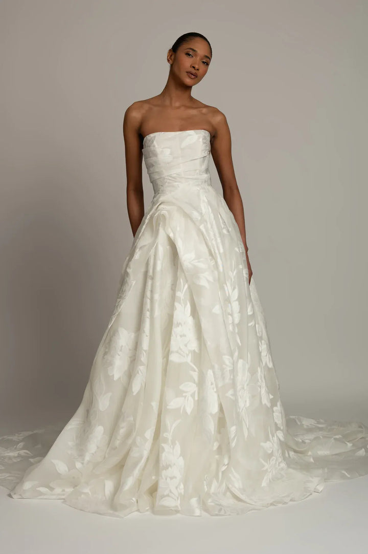 A woman modeling a strapless white bridal gown with a structured bodice and a full, flowing skirt featuring tonal floral jacquard texture, photographed against a plain light gray studio background.