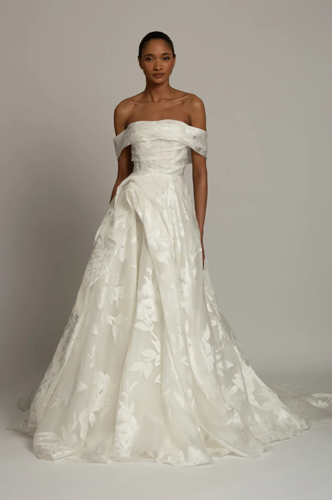 An off-the-shoulder white ball gown with a structured bodice and a full, textured skirt, modeled against a neutral light gray studio background.