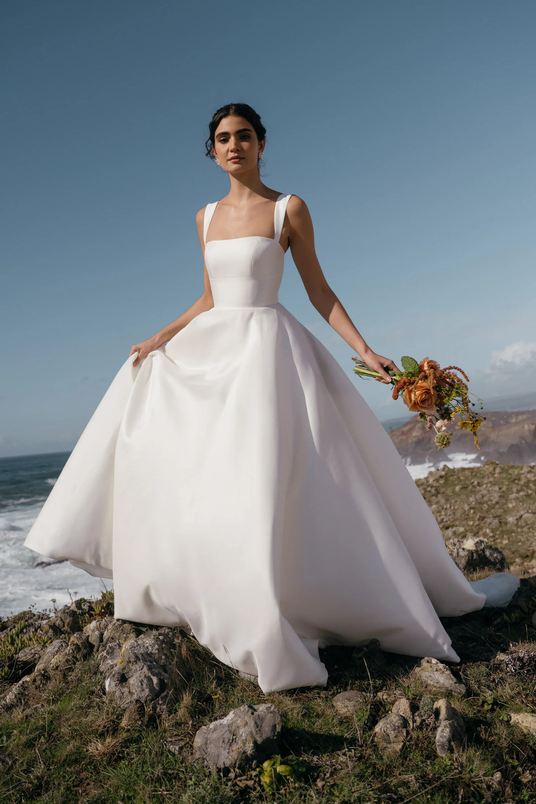 A woman wearing a sleeveless white wedding gown with a structured bodice and full skirt stands on rocky coastal ground, holding a bouquet of flowers against a blue sky and ocean background.