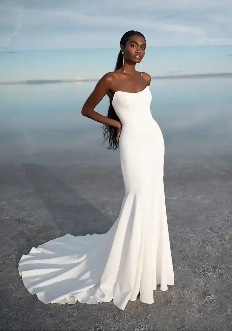 A woman modeling a strapless white mermaid-style gown with a fitted bodice, flared skirt, and long train, standing outdoors against a calm reflective water surface under a pale sky.
