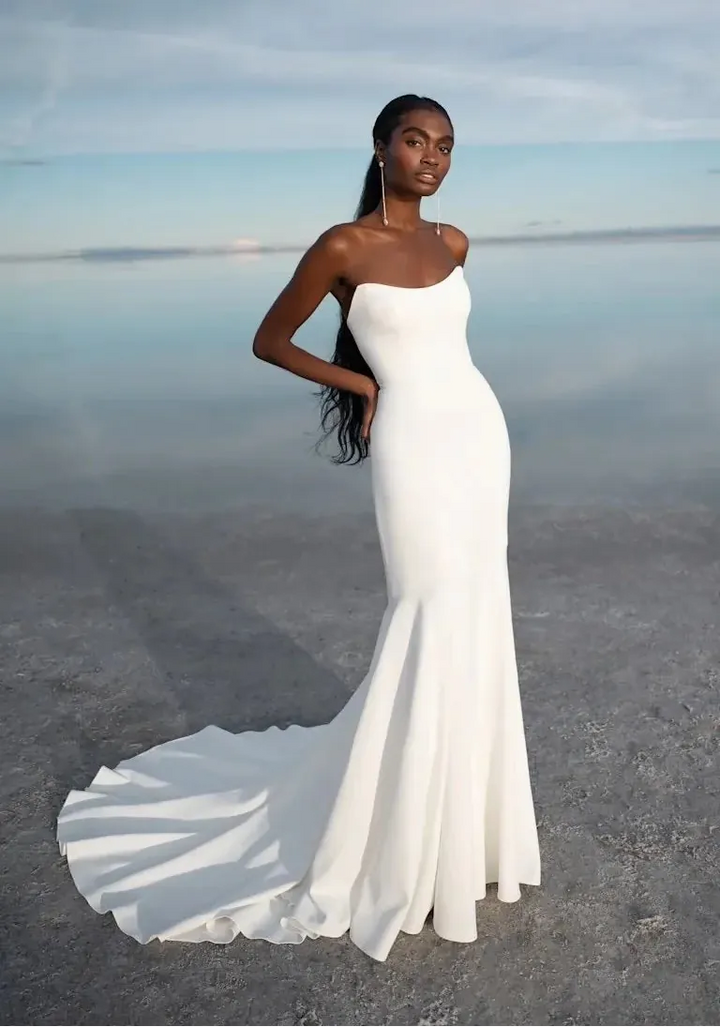 A woman modeling a strapless white mermaid-style gown with a fitted bodice, flared skirt, and long train, standing outdoors against a calm reflective water surface under a pale sky.