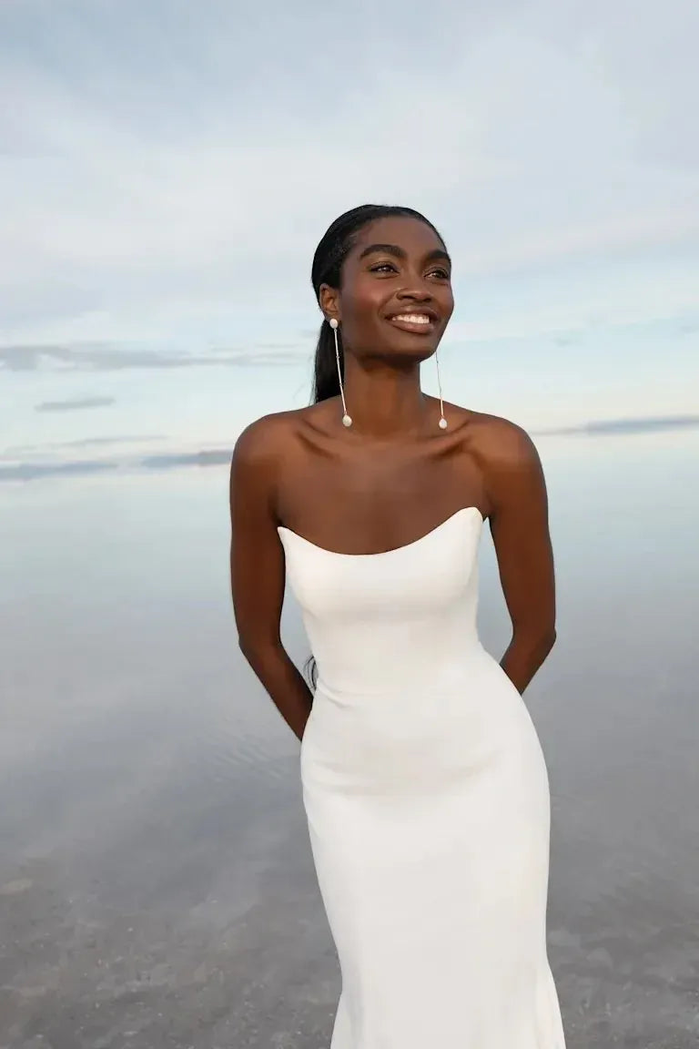 A woman wearing a strapless white mermaid-style dress stands outdoors against a calm water and sky background, smiling and looking upward.