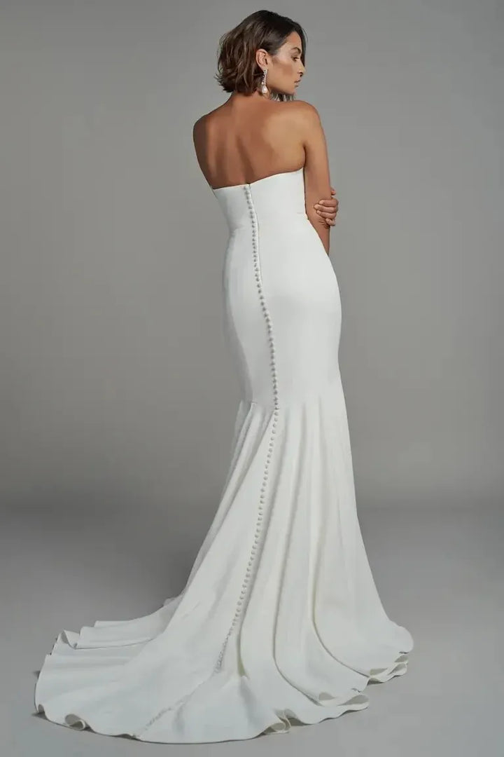 A woman modeling a strapless white mermaid-style wedding gown from the back, showing a fitted silhouette, a long train, and a vertical line of embellished back studs against a plain gray studio background.