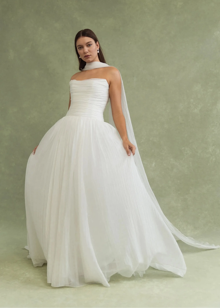 A woman modeling a strapless white wedding gown with a full tulle skirt and a long sheer veil, posed against a textured sage green studio backdrop.