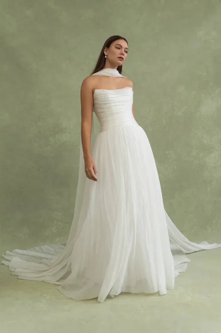 A woman modeling a strapless white wedding gown with a fitted ruched bodice and a flowing tulle skirt with a long train, posed against a textured sage green studio backdrop.