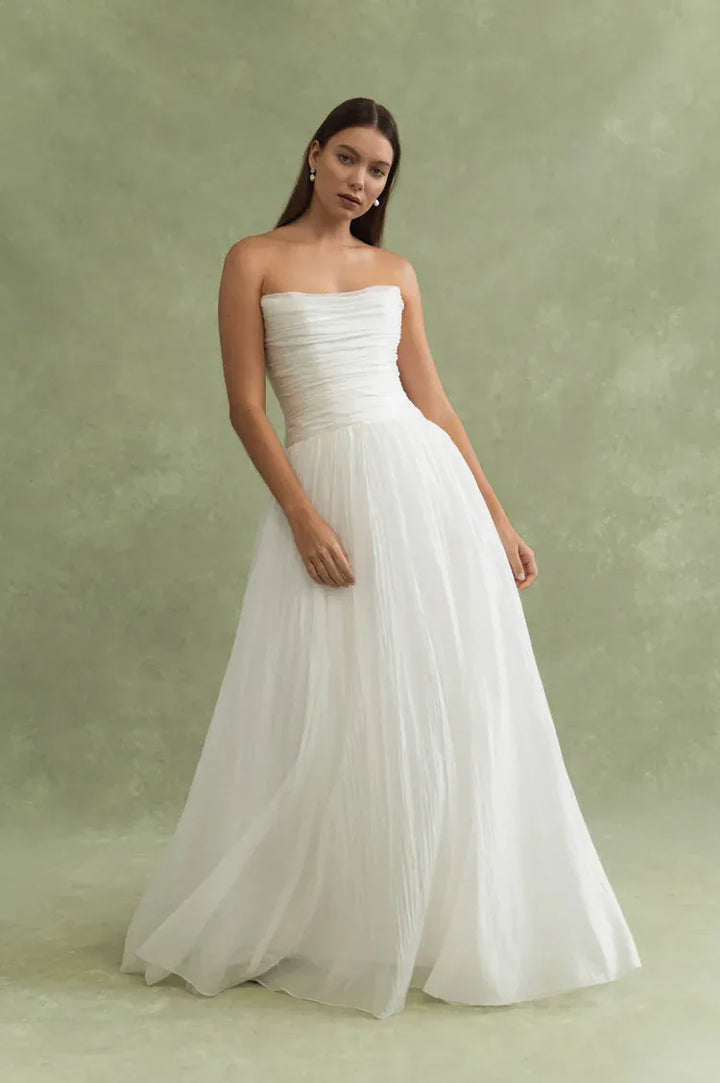 A woman modeling a strapless white wedding gown with a ruched bodice and a full, flowing tulle skirt against a textured sage green studio backdrop.