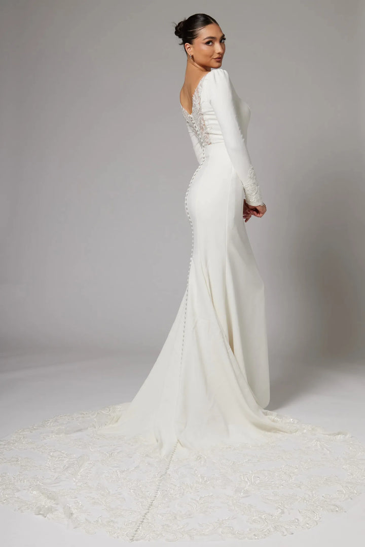 A woman modeling a long-sleeve white wedding gown with a fitted silhouette, lace-embellished back, and a long lace train against a light gray studio background.