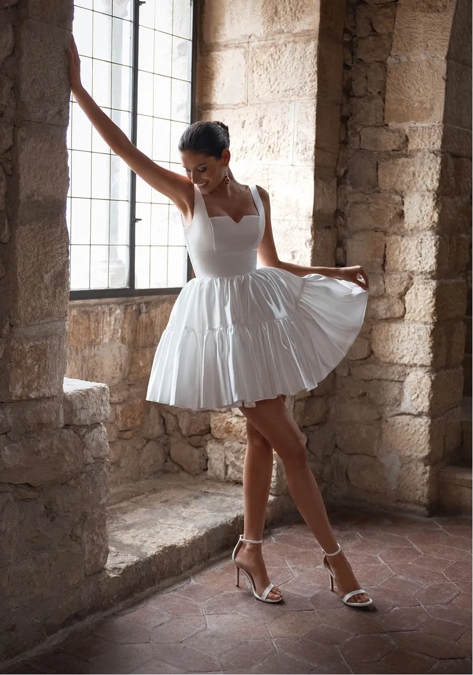 Woman in a white dress standing in a stone-walled room with large windows.
