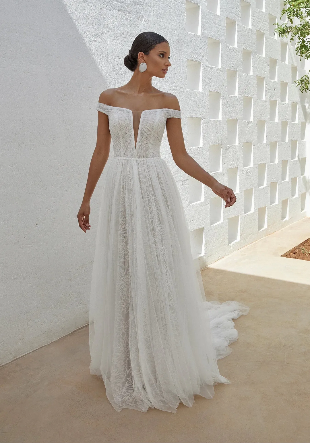 Woman in a white lace wedding dress standing against a textured white wall.