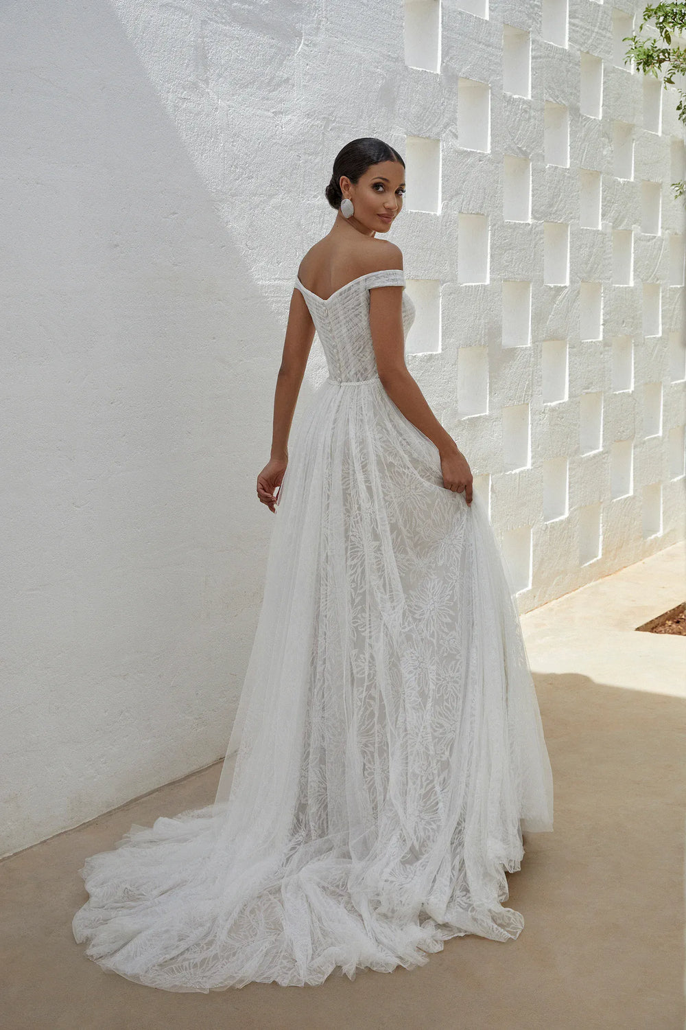 Woman in a white lace wedding dress standing against a textured white wall.