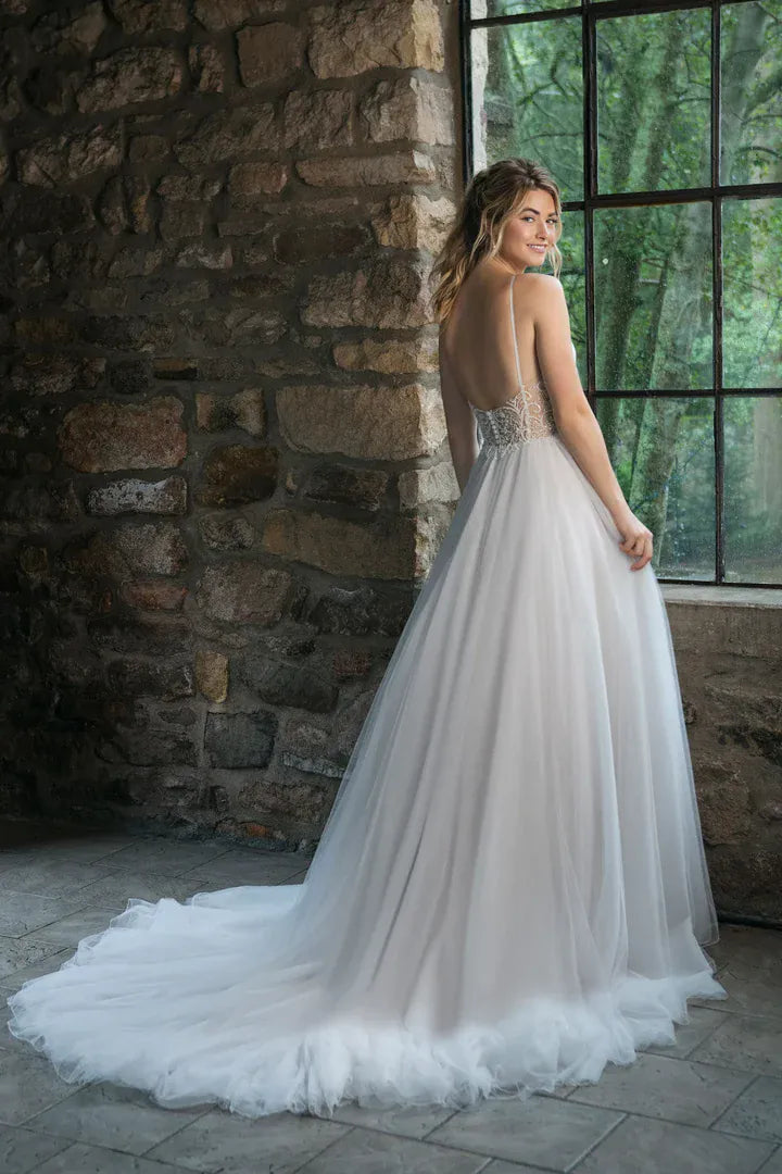 A woman wearing a voluminous white wedding gown stands in a stone interior near a large window, photographed from the back to show the back of the dress and train.