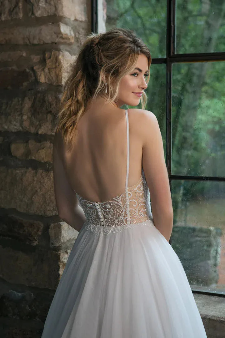 A woman wearing a white wedding dress with a sheer lace back and tulle skirt, standing indoors beside a stone wall and large window.