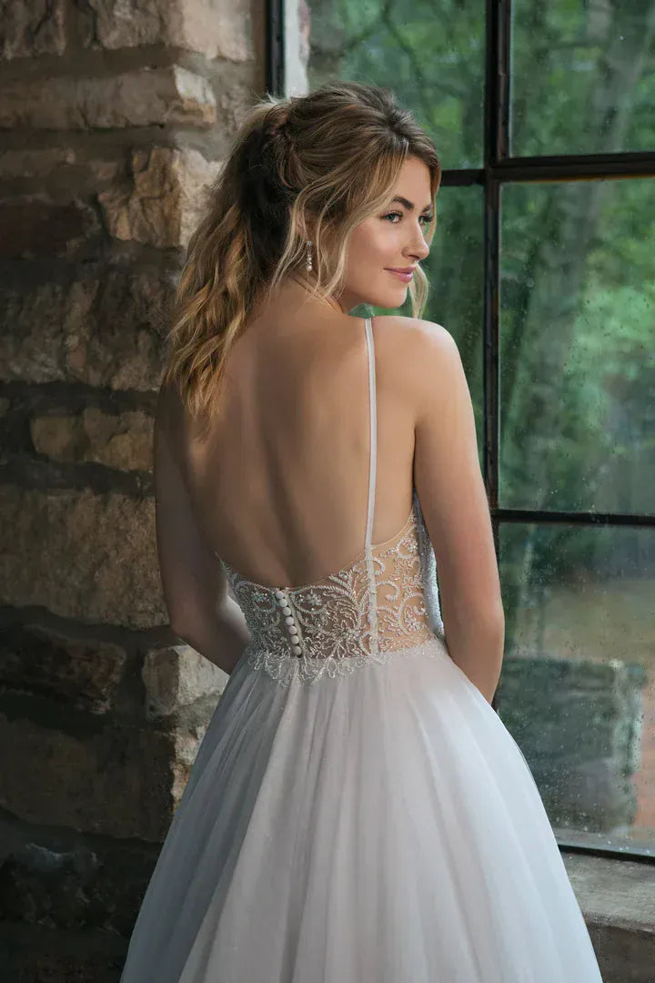 A woman wearing a white wedding dress with a sheer lace back and tulle skirt, standing indoors beside a stone wall and large window.