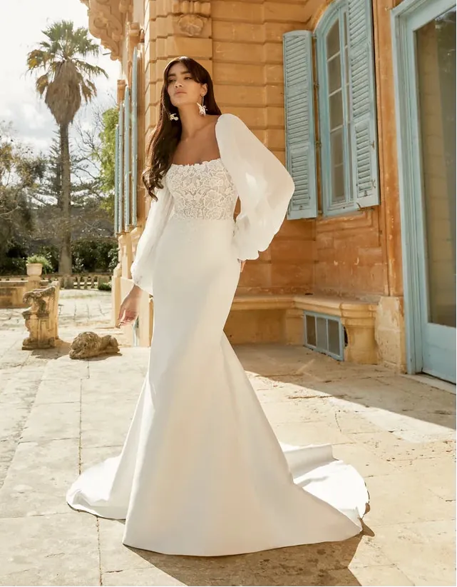 A woman modeling a white wedding gown with a lace bodice, square neckline, and dramatic puffed sleeves, standing outdoors in front of a warm stone building with blue shutters.