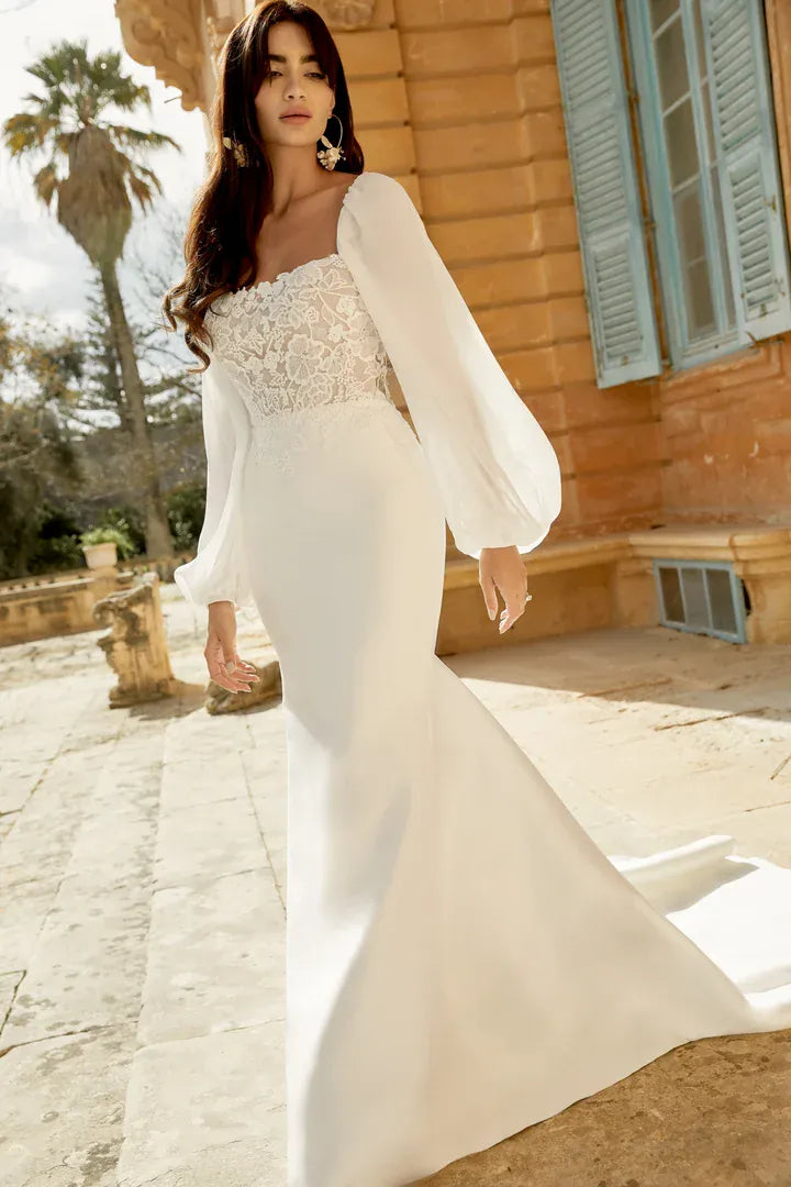 A woman wearing a white wedding gown with a lace-embellished sweetheart bodice, sheer balloon sleeves, and a fitted mermaid-style skirt, standing outdoors in front of a warm stone building with blue shutters.