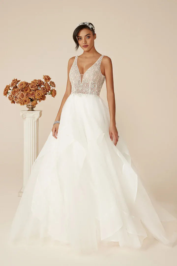 A woman models a white wedding gown with a beaded lace bodice, deep V-neckline, and a voluminous layered tulle skirt. She is standing in a studio setting with a beige background, next to a white pillar with dried orange-brown flowers.