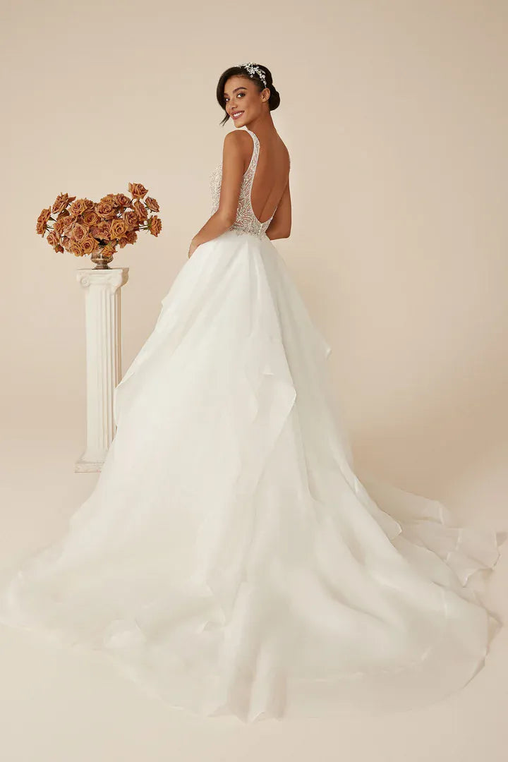 A woman modeling a white wedding dress from the back, standing in a studio with a warm beige background. The dress has a fitted lace bodice, a low open back, and a full layered tulle skirt with a long train.