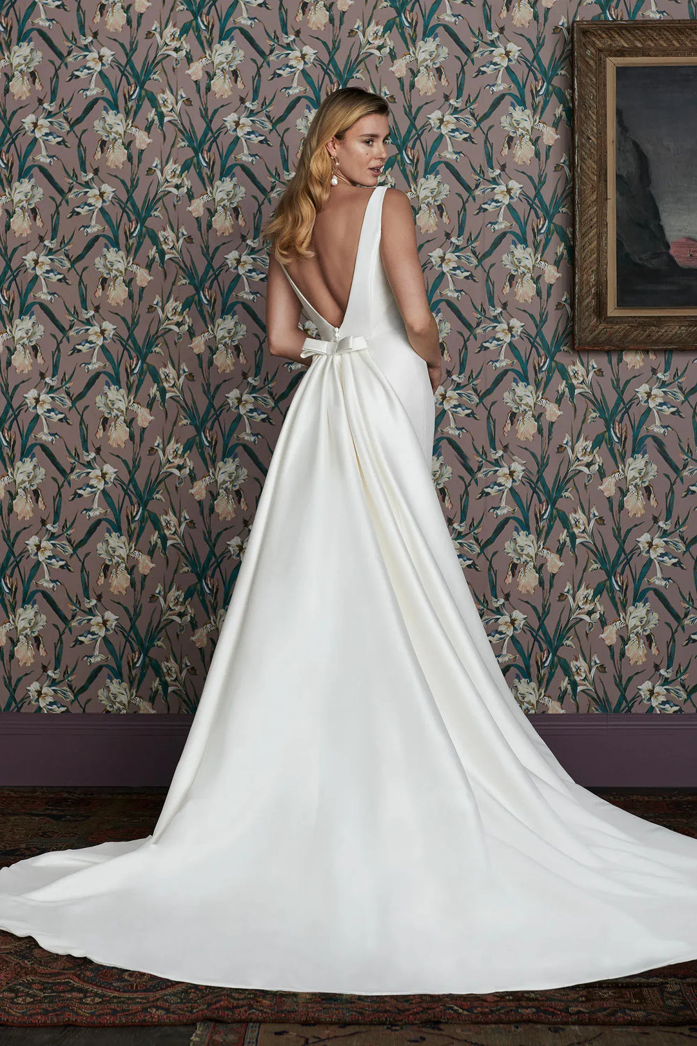 Woman in a white wedding dress with a long train standing in front of floral wallpaper.