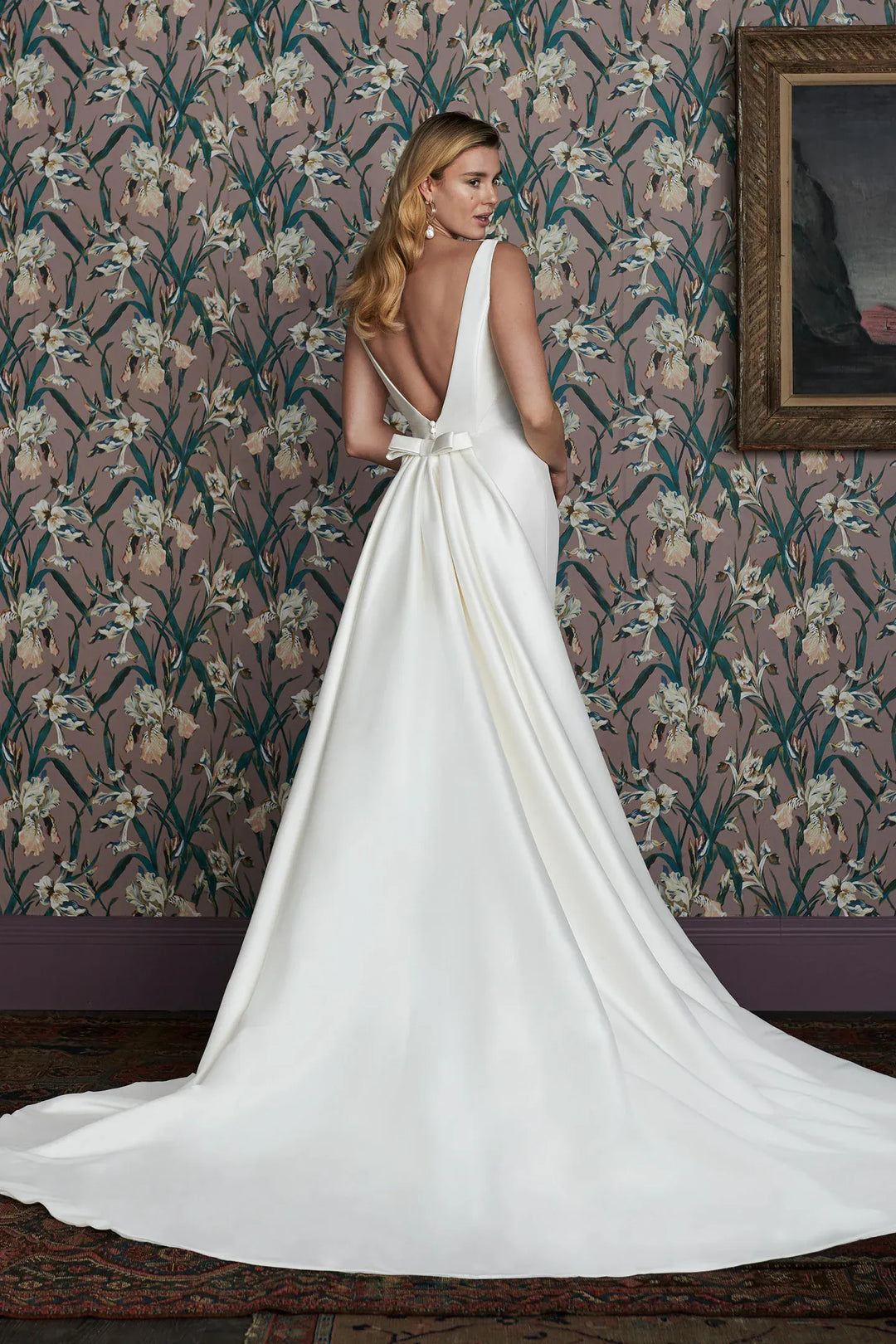 Woman in a white wedding dress with a long train standing in front of floral wallpaper.