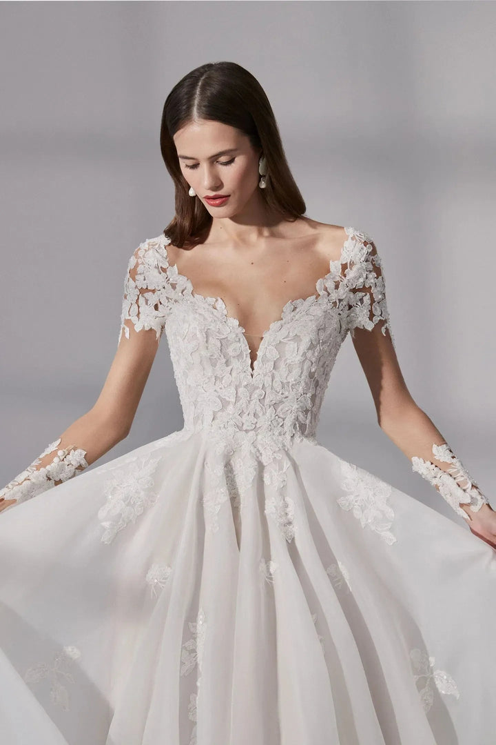 A woman wearing a white lace wedding dress with a deep V-neckline, short lace sleeves, and a full tulle skirt, posed against a soft gray studio background.